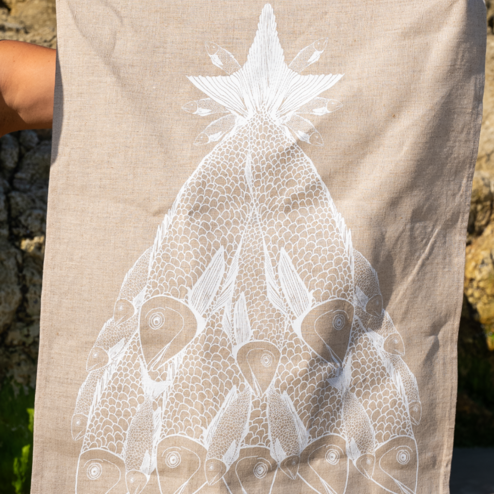 Merry Fishmas linen tea towel held open outdoors, showing the full white fish design on natural flax