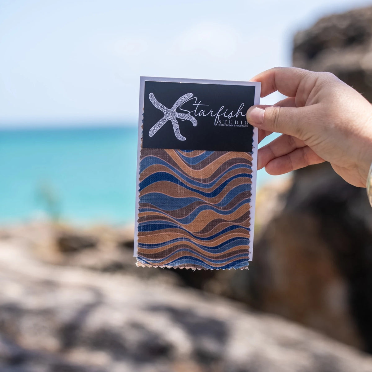Straddie Stripe Mariner linen fabric swatch in navy blue and ochre held beside the ocean