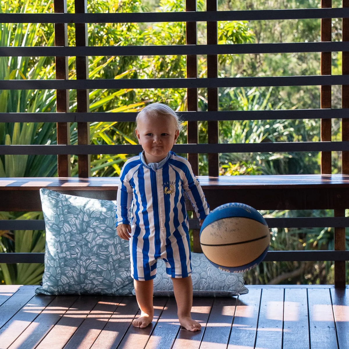 Toddler standing on a timber deck with a basketball, styled in front of Frenchmans colourway cushions featuring the Pipi and Growth Rings patterns.