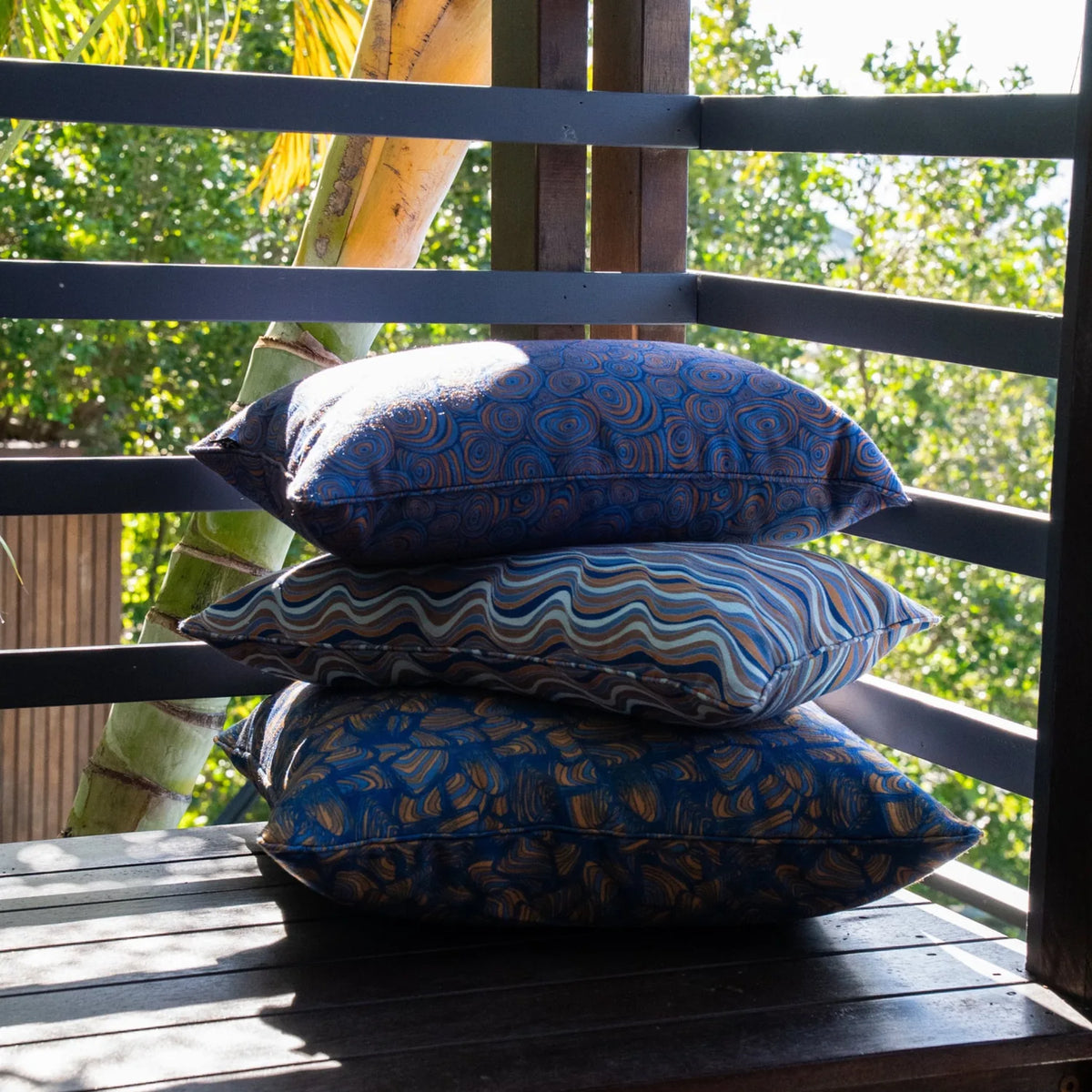 Stack of three Mariner cushions on a timber bench, showing Growth Rings, Straddie Stripe and Pipi prints.