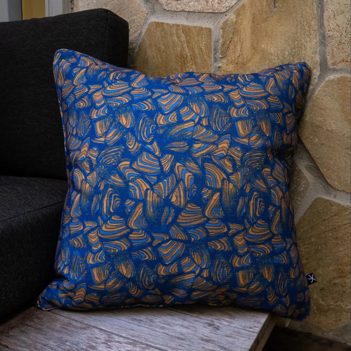 Square Pipi Mariner cushion with blue and tan abstract print styled against a stone wall.