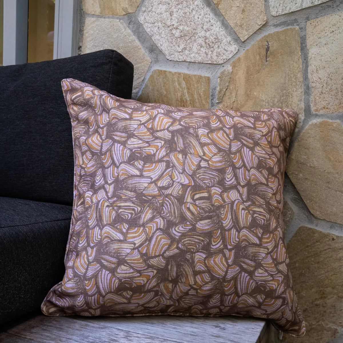 Claytons Pipi outdoor indoor cushion cover in a square size styled on an outdoor chair beside a stone wall.