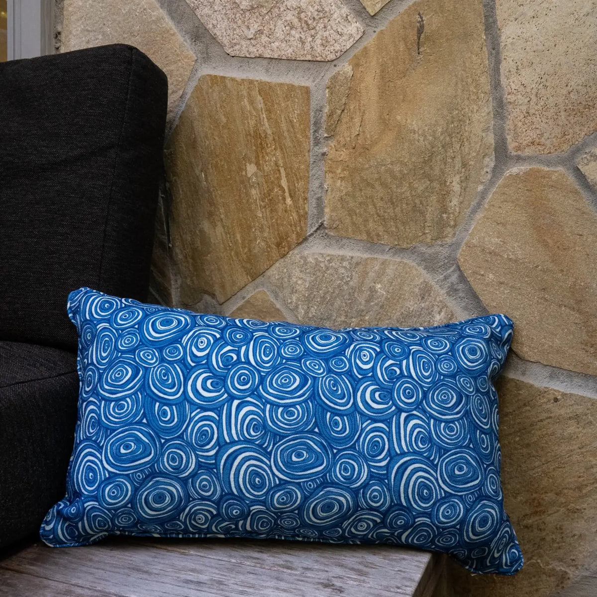 Ocean Growth Rings outdoor indoor cushion cover in a rectangular lumbar size styled on an outdoor chair beside a stone wall.
