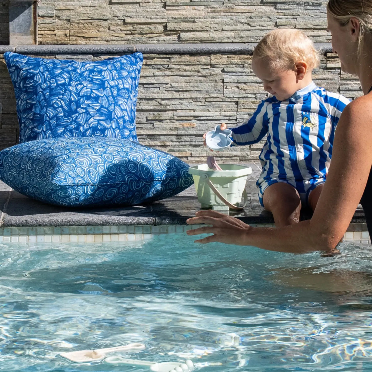 Ocean Growth Rings outdoor indoor cushion cover in a square size styled on an outdoor chair beside a stone wall.