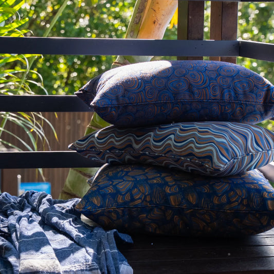 Stack of three patterned outdoor cushions styled on a timber deck with a blue striped throw.